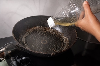 Una mano sostiene una botella de plástico transparente y vierte unas gotas de aceite en una sartén negra colocada sobre una estufa.