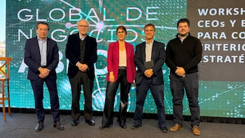 Cinco personas de pie en un escenario posan ante una pantalla digital con fondo verde y texto en blanco