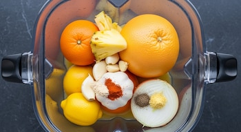 Una licuadora transparente contiene naranjas enteras, un pomelo, limones, trozos de piña, dientes de ajo, media cebolla y montones de especias en polvo.