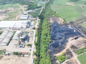 Vista aérea de una planta industrial junto a una zona de terreno oscuro, separadas por una hilera de árboles, con campos verdes y caminos de tierra