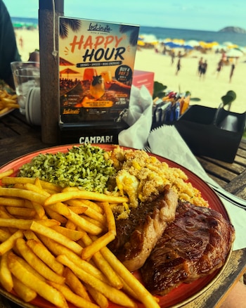 Un plato con carne asada, papas fritas, arroz verde y farofa sobre una mesa de madera, con un cartel publicitario y una playa de fondo