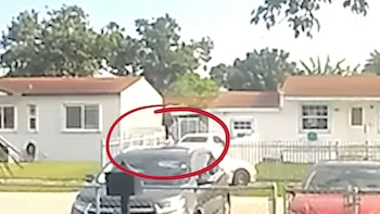 Un círculo rojo encierra a una persona y un automóvil blanco. Se ven otros dos automóviles, casas con tejados naranjas y árboles verdes