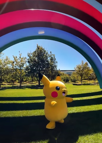 Una botarga amarilla de Pikachu sobre el césped verde en un parque con árboles y una estructura en forma de arco multicolor