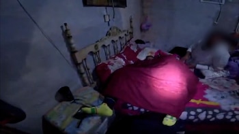 Habitación oscura con cama, sábanas estampadas, persona acostada cubierta por manta roja, otra persona sentada, luz de linterna iluminando una zona