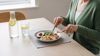 Manos de una persona cortando pollo con cuchillo y tenedor en un plato blanco, acompañado de guarnición, sobre una mesa de madera clara.