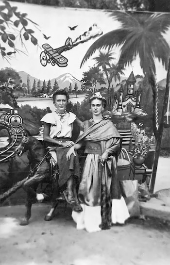 Dos mujeres, una sentada sobre un caballo de utilería y otra de pie, posan frente a un telón pintado con montañas, árboles, un tren, un avión y un castillo