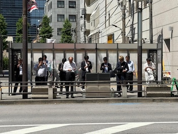 Grupo de personas, mayormente hombres en trajes de negocios, de pie en un área de fumadores designada al aire libre en una calle de Tokio. Algunos fuman, otros miran sus teléfonos