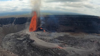 Vista aérea de una erupción volcánica en el cráter de Kīlauea, con lava naranja brillante elevándose en una fuente y fluyendo sobre terreno rocoso. Humo blanco