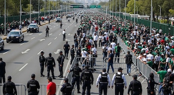 Una avenida con vehículos policiales, numerosos agentes de policía y una gran multitud de personas. Hay vallas metálicas que separan a la gente de la vía.