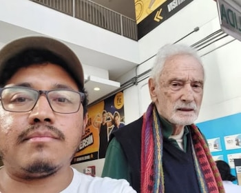 Selfie de un joven con gafas y gorra junto a Guillermo Giacosa, un hombre mayor con cabello y barba blancos, y una bufanda colorida, en un interior