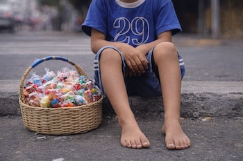 Los pies y las piernas de un niño descalzo sentado en una acera, con una camiseta azul, junto a una canasta llena de dulces envueltos.