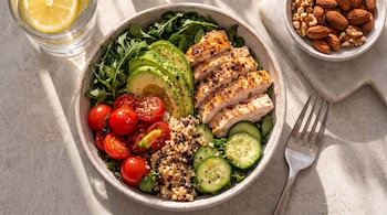 Vista aérea de un bol de comida saludable con pollo a la parrilla en rodajas, aguacate, tomates cherry, quinoa, pepino y rúcula, con agua de limón y frutos secos.