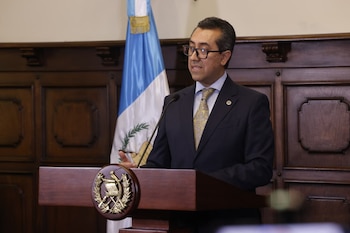 Hombre con traje y gafas habla en un podio. Bandera de Guatemala a la izquierda, pantalla digital con documentos a la derecha. Fondo con paneles de madera