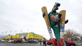 Estatua gigante de dinosaurio verde con botas de vaquero, sosteniendo postes. Fondo: edificio amarillo "The Big Texan" y coches en el aparcamiento
