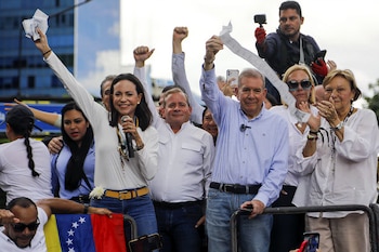María Corina Machado y Edmundo González Urrutia sonríen y levantan papeles en un mitin, rodeados de gente. Se ve una bandera venezolana