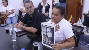 Una mujer en camisa blanca habla en una mesa mientras sostiene un póster con el rostro de una joven; un hombre de barba observa a su lado