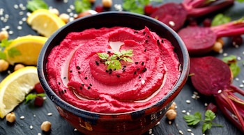 Cuenco de hummus de remolacha, perejil, semillas de sésamo negras. Alrededor: rodajas de limón, garbanzos, sésamo y remolachas cortadas.