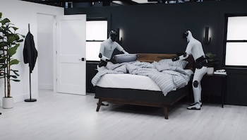 Dos robots humanoides blancos y negros están en un dormitorio moderno; uno ajusta un cojín en una cama con ropa de cama gris, el otro está de pie cerca