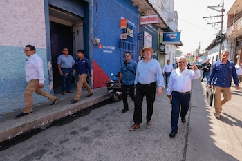 Grupo de personas, incluyendo a Joaquín Barnoya, camina por una carretera de asfalto, algunos visten ropa tradicional. Hay árboles y un puente al fondo