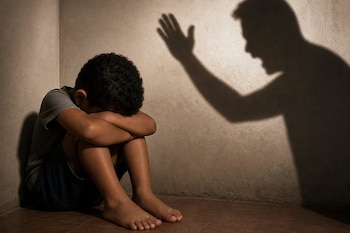 Un niño sentado en el suelo de una esquina con la cabeza sobre las rodillas, junto a la sombra de una persona con la mano alzada en la pared.