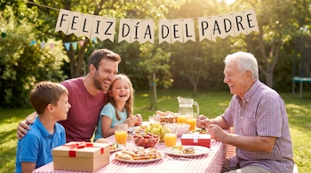 Un hombre, un anciano, un niño y una niña sentados alrededor de una mesa de picnic con comida y regalos, riendo bajo un cartel de 'Feliz Día del Padre'.
