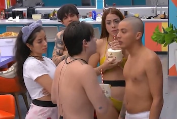 Un grupo de cinco jóvenes en una cocina de tonos vibrantes; tres están sin camiseta o en bikini, y una mujer le da de beber de un coco a un hombre con una pajita