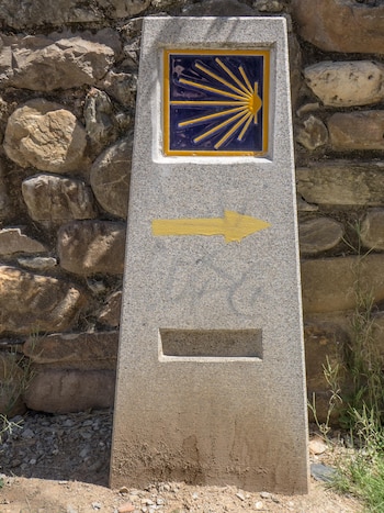 Marcador de granito del Camino de Santiago con azulejo azul y amarillo de concha de vieira y una flecha amarilla a la derecha, sobre un muro de piedra