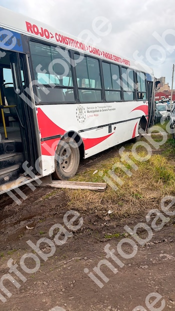 Un colectivo blanco y rojo varado en terreno embarrado con pasto seco, sin una rueda delantera y apoyado en tablas de madera