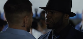 50 Cent con gorra de béisbol negra con el logo de los Yankees, camisa estampada azul y blanca, y pendientes, posa de frente sobre un fondo gris oscuro