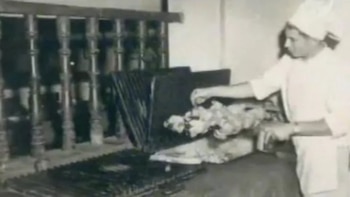 Hombre vestido de cocinero con gorro y uniforme blanco manipulando pollos en un horno giratorio con rejillas metálicas y un barandal de madera al fondo