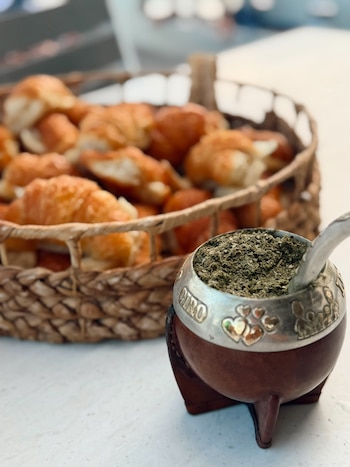 Primer plano de un mate de calabaza y metal con yerba y bombilla, junto a una canasta de mimbre con múltiples croissants dorados