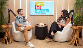 Josh Horowitz y Adria Arjona están sentados en sillones frente a frente con micrófonos, ante una pantalla del programa Happy Sad Confused
