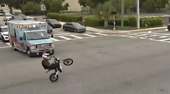 Motociclista con casco blanco haciendo una acrobacia sobre una rueda. Ambulancia con luces, coches, y cruce peatonal en la calle