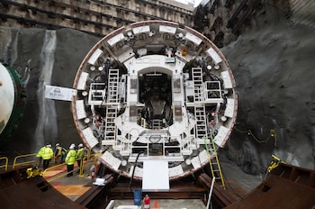 Una enorme tuneladora circular blanca junto a una pared de roca y varios trabajadores con chalecos amarillos de alta visibilidad