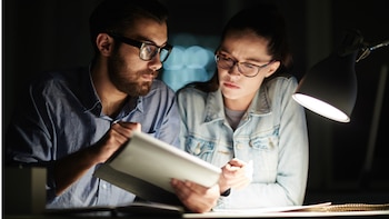 Un hombre y una mujer con lentes observan un documento sobre un escritorio iluminado por una lámpara en un espacio oscuro