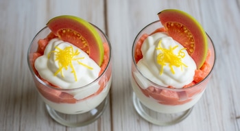 Toma cenital de dos copas con postre de guayaba rosa y capas blancas de yogur, decoradas con una rodaja de guayaba y ralladura de limón amarillo.