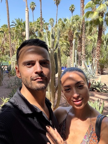 Selfie de Rita Baroud y un hombre en un jardín con palmeras y múltiples cactus. Ambos miran a la cámara bajo la luz del sol