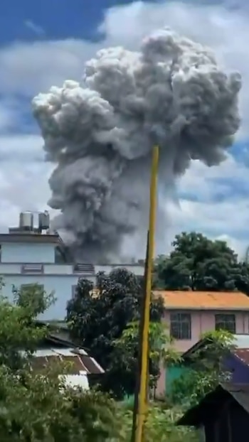 Una masiva columna de humo gris oscuro se eleva hacia el cielo, vista desde detrás de edificios de dos pisos y árboles verdes en un día soleado