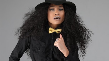 Mujer con cabello rizado, sombrero negro, pajarita amarilla con diseño de abejas, camisa negra, labios plateados y un anillo en la mano derecha