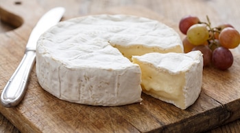 Un queso camembert con una porción cortada, un cuchillo metálico y un racimo de uvas sobre una tabla de madera.