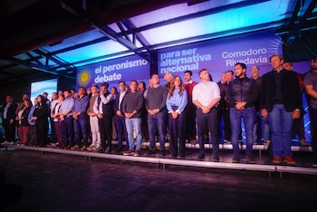 Un grupo de personas permanece de pie sobre una tarima frente a un fondo azul con inscripciones y focos de colores