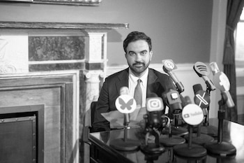 Un hombre con barba y traje, identificado como Mamdani, sentado frente a una mesa con varios micrófonos de redes sociales en blanco y negro