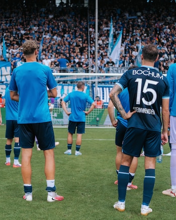 Jugadores de fútbol vistos de espalda en el campo mientras observan una tribuna con espectadores sosteniendo banderas azules y blancas