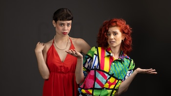Dos mujeres posan sobre un fondo oscuro: una viste vestido rojo y collar de perlas, y la otra lleva una camisa multicolor y cabello teñido de rojo