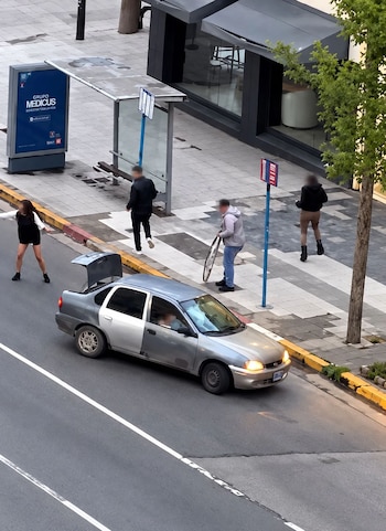 Vista desde arriba de un automóvil gris con el maletero abierto en una calle y varios peatones con caras difuminadas sobre la acera