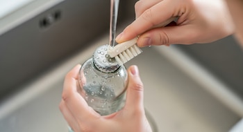Primer plano de manos limpiando la tapa metálica de una botella de vidrio con un cepillo bajo un chorro de agua. Gotas de agua visibles en la botella y el fregadero.