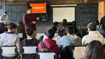 Un hombre de pie habla frente a alumnos sentados en un aula que tiene pizarras y una pantalla con el texto Educación Financiera
