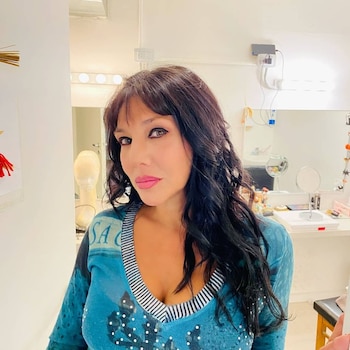 Una mujer de cabello oscuro y suéter azul posa frente a la cámara en una sala con espejos y luces