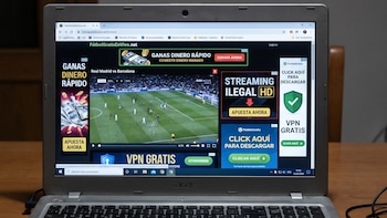 Un ordenador portátil sobre una mesa de madera con su pantalla encendida mostrando un partido de fútbol, una página web con anuncios, una taza y un ratón.