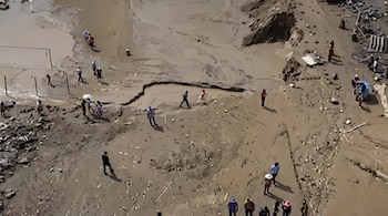 Vista aérea de personas caminando sobre un terreno de barro y arena con charcos de agua, huellas, piedras y una valla metálica.